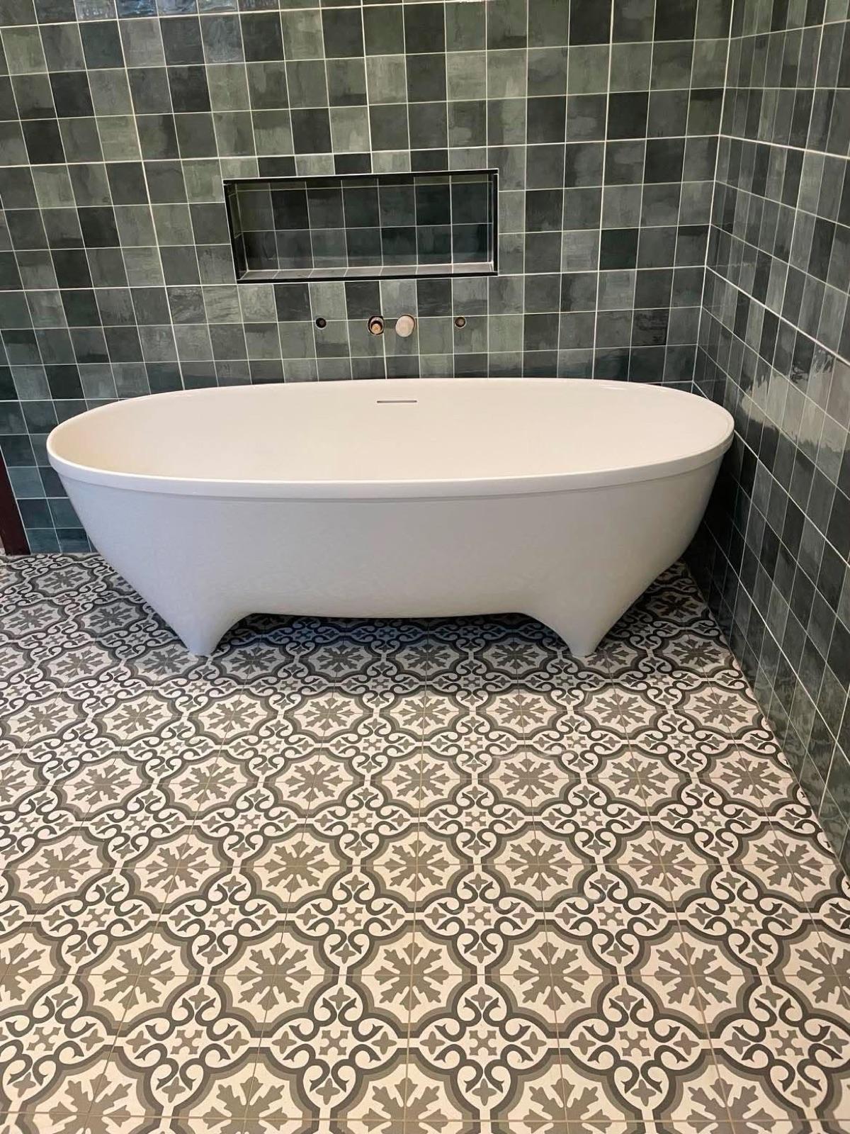 A finished bathroom with a white freestanding bath against green mosaic tiles and a patterned floor.